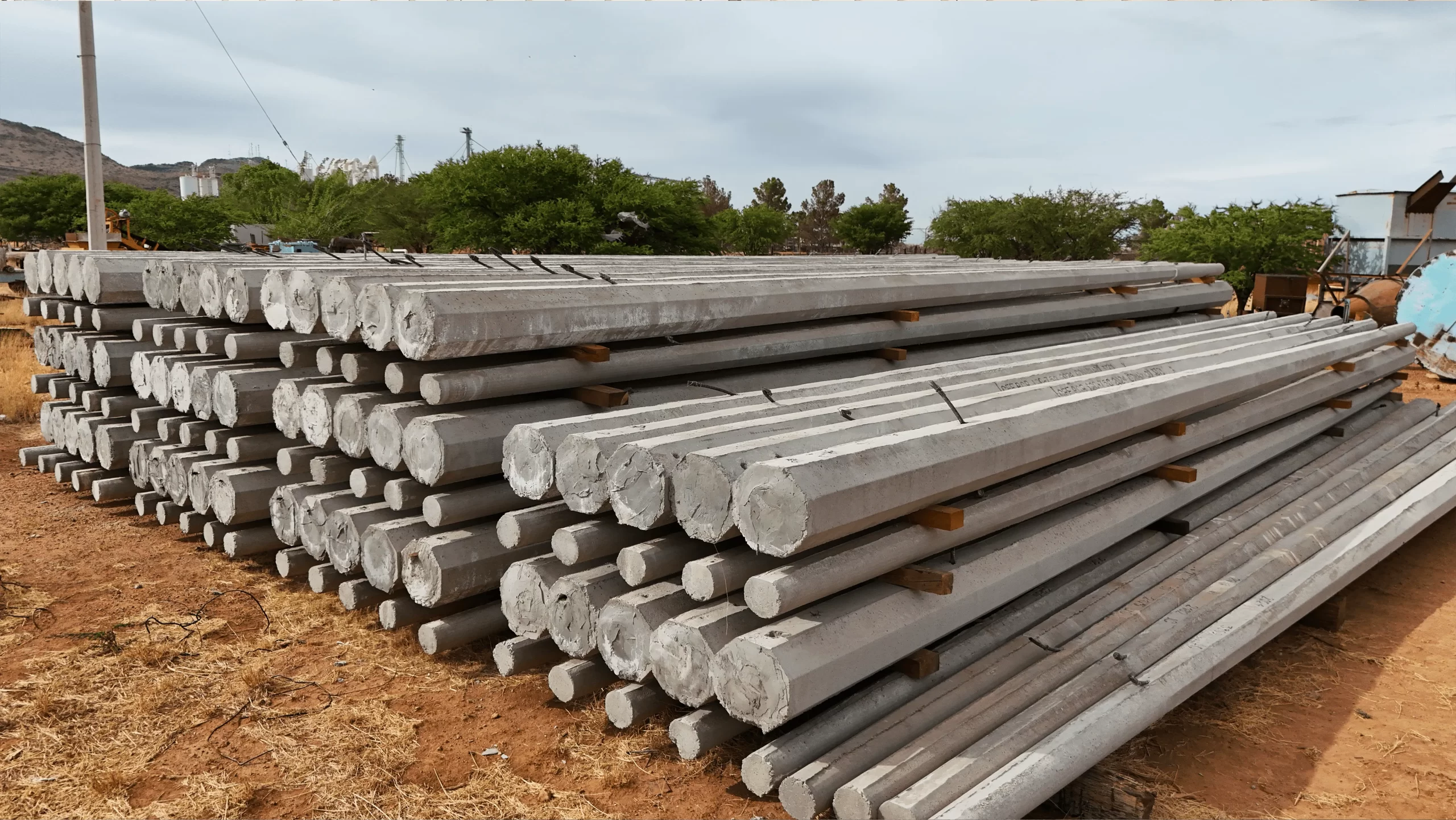 Grupo de postes de concreto prefabricados acomodados en la planta de GP Prefabricados, con fondo de nubes y árboles, líderes en soluciones para redes eléctricas en el norte de México.